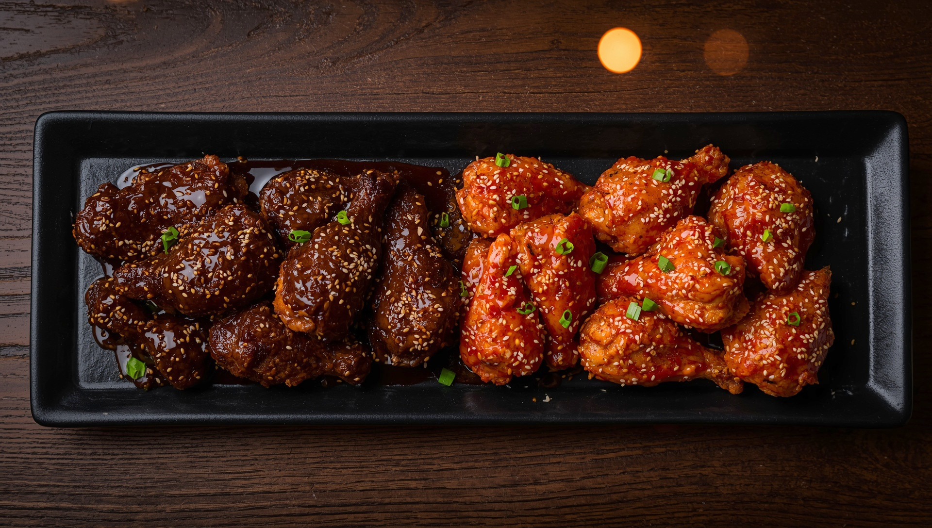 A whole chicken on a long black platter, one half glazed dark soy garlic brown and the other half bright red gochujang.